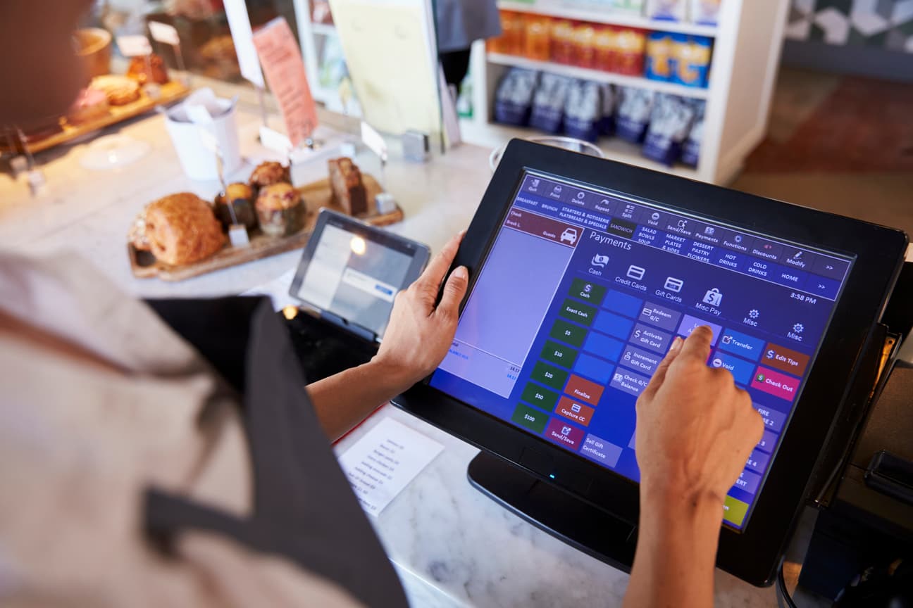 Hands using a touch-screen point-of-sale system at a bakery counter with pastries nearby.