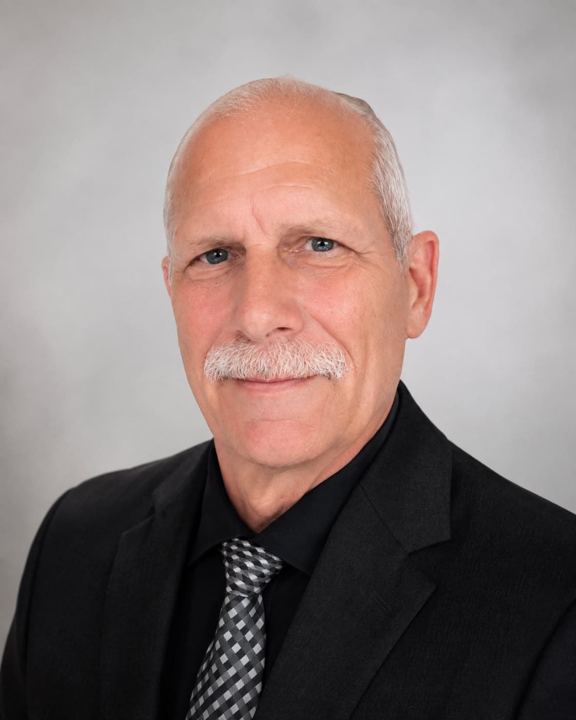 Professional headshot of a man with a white mustache, black suit, and checkered tie.