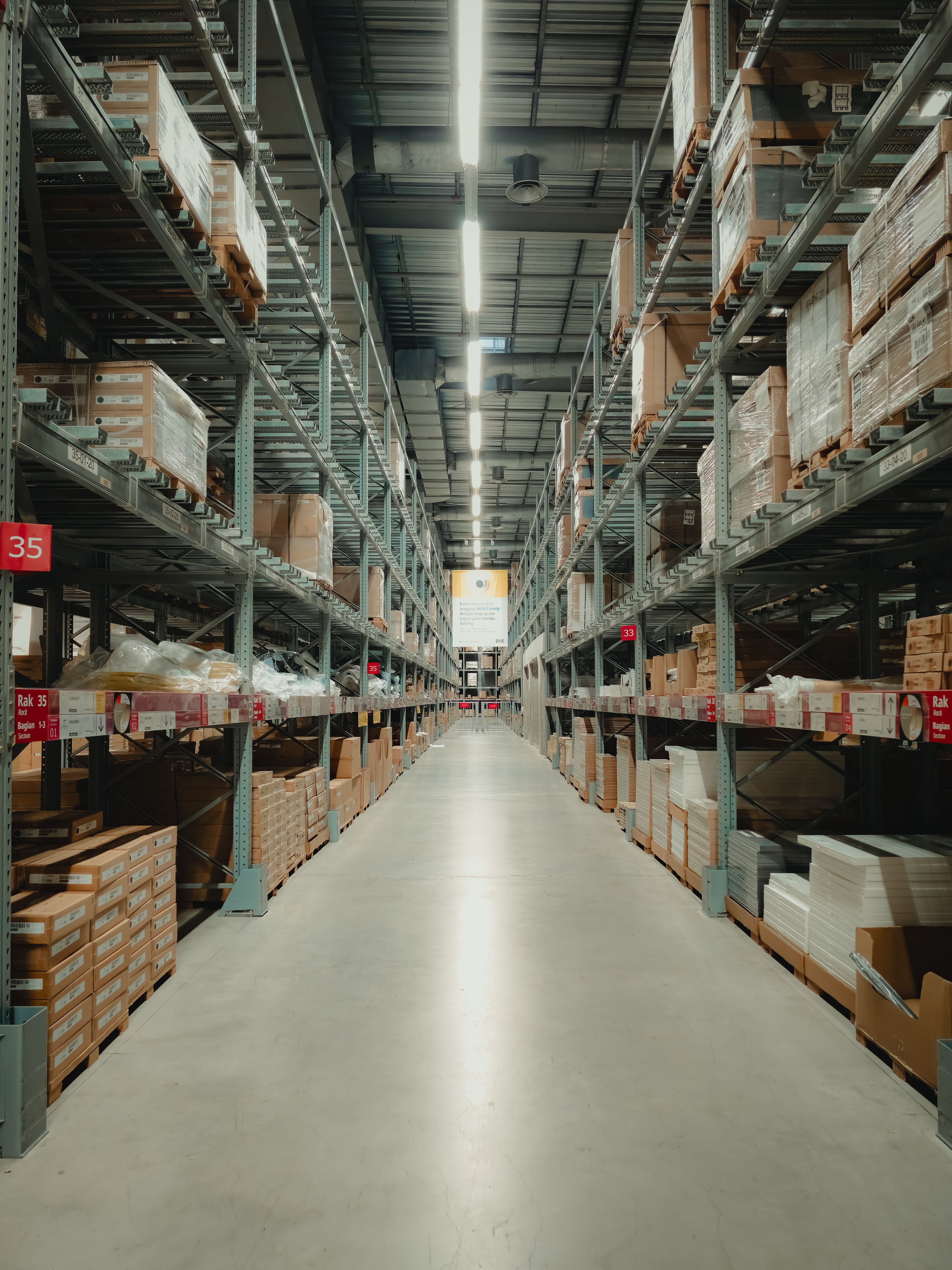 High metal shelves filled with cardboard boxes line a long, brightly lit warehouse aisle.