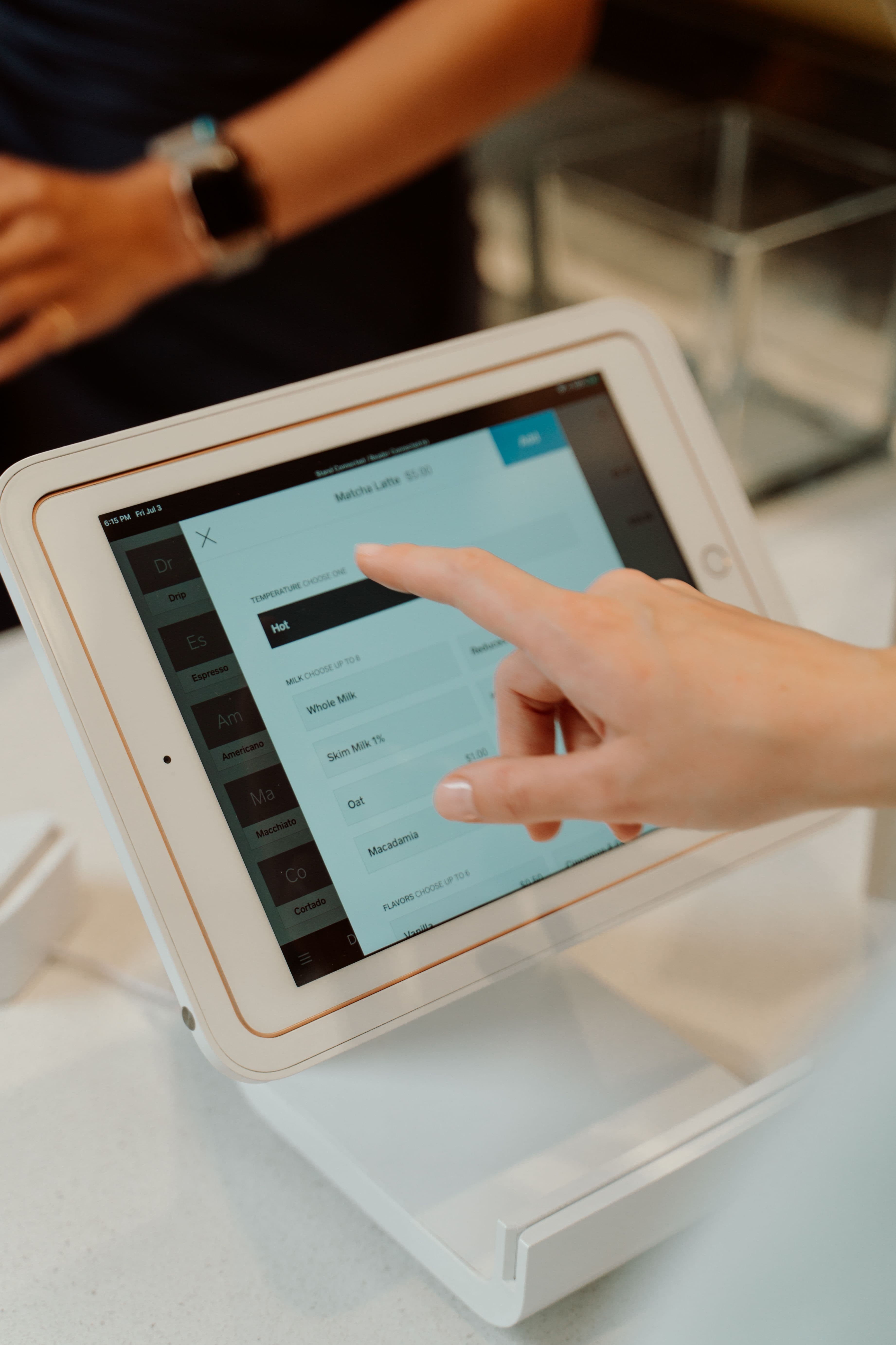 A hand touches a white tablet screen to customize a matcha latte order at checkout.