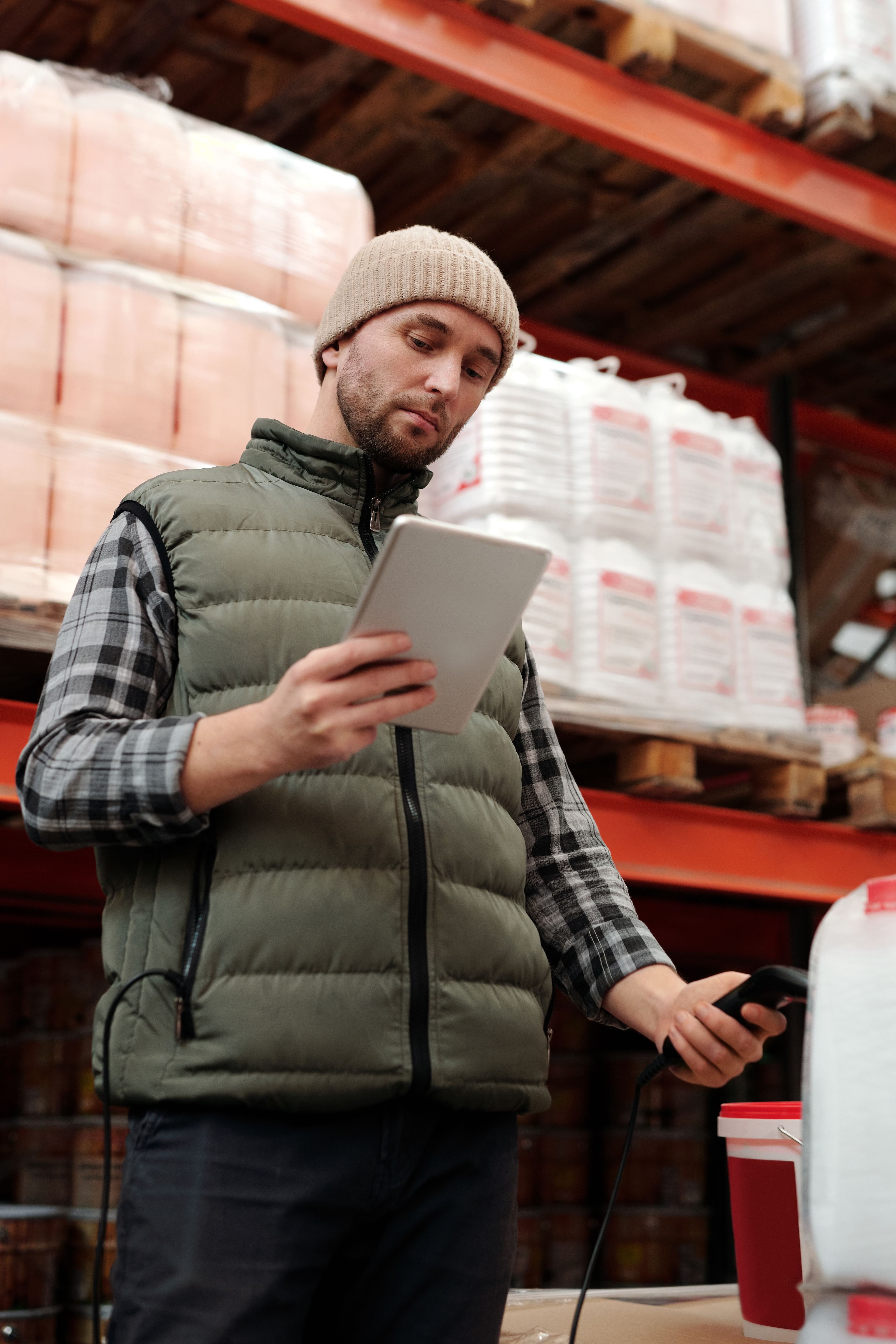 A warehouse worker in a puffer vest and beanie uses a tablet and barcode scanner.