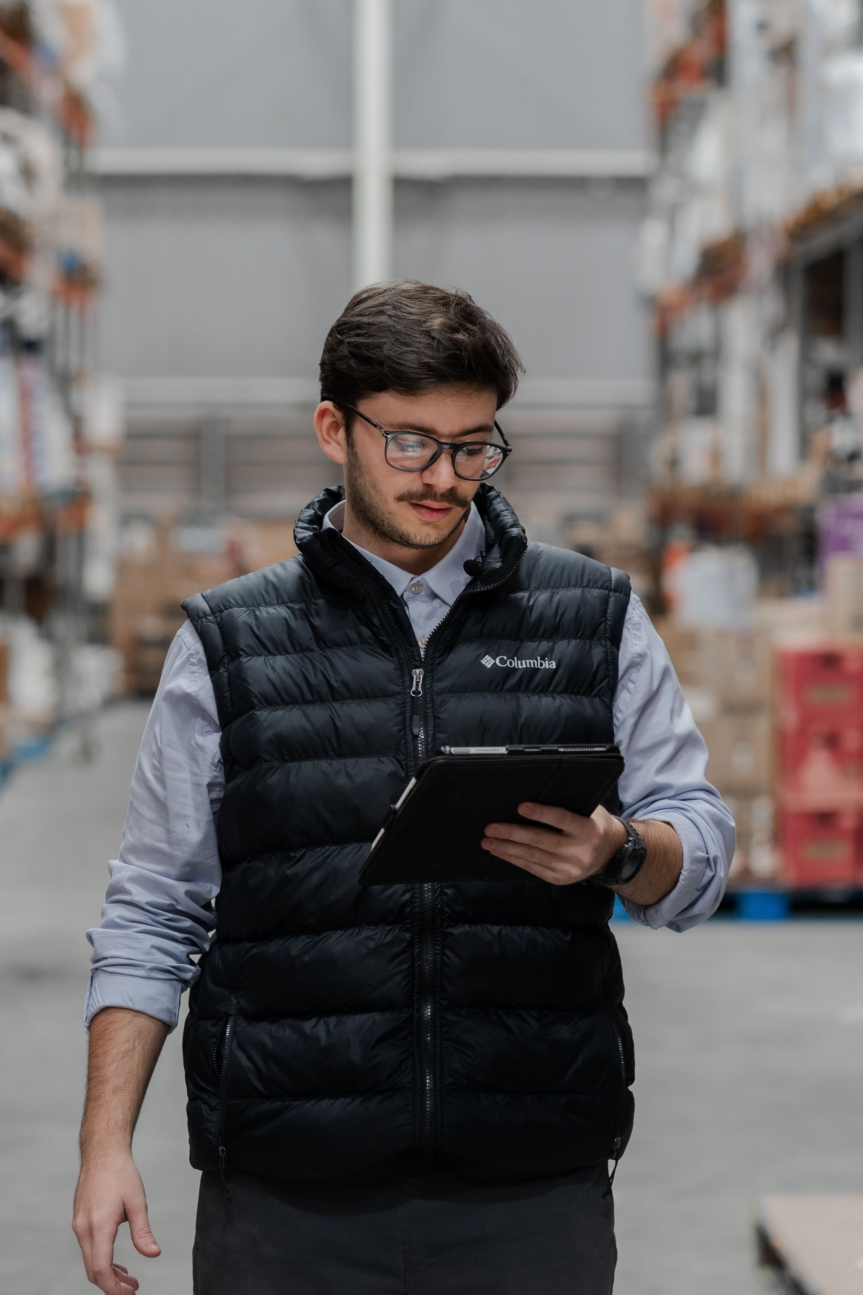 Man in a black puffer vest and glasses using a tablet in a warehouse.