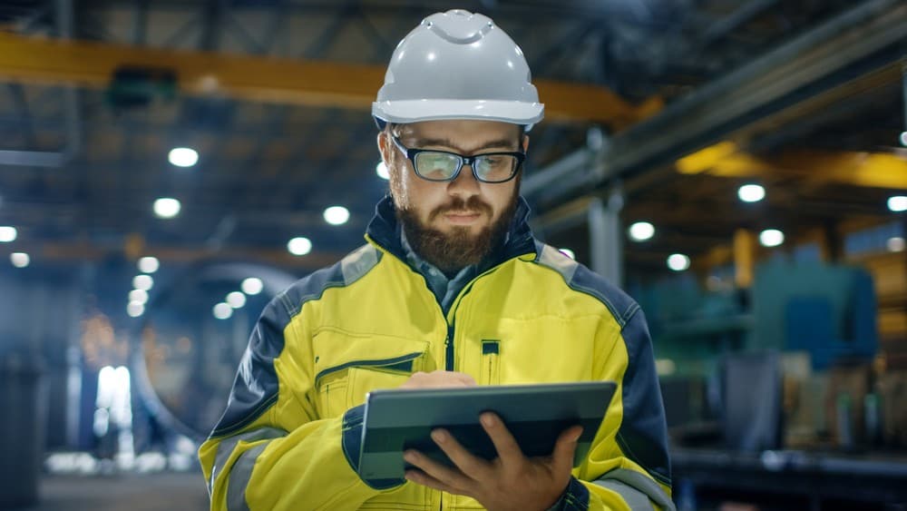 Industrial worker in a hard hat and safety jacket using a tablet in a factory.