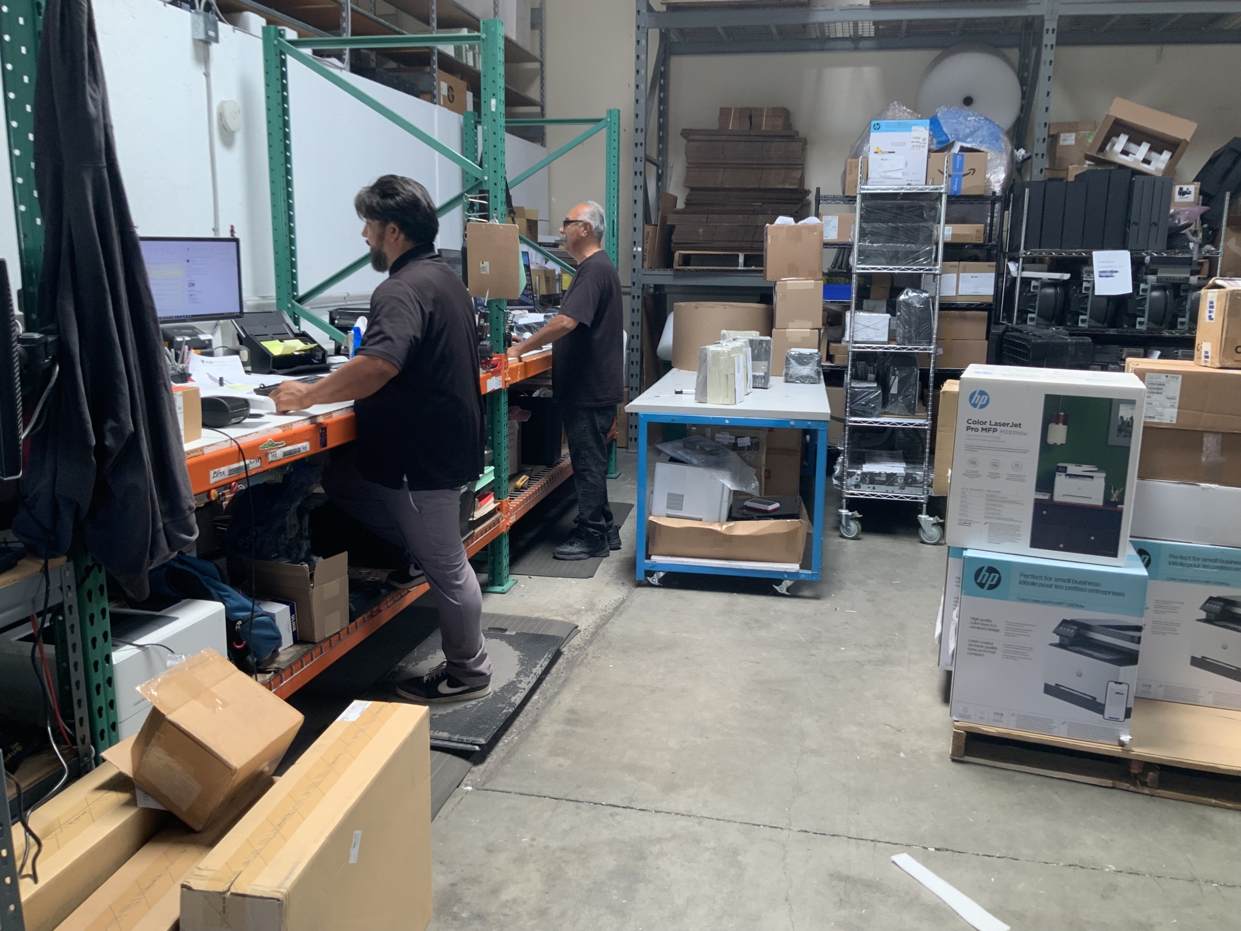 Two men work at computers in a warehouse room full of equipment and other inventory.
